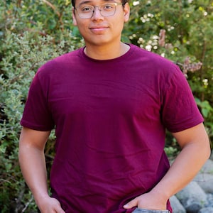 May include: A solid burgundy short-sleeved t-shirt with a crew neckline. The shirt is made of a plain fabric and is worn with grey denim jeans. The design is simple and features a standard fit with short sleeves that reach the mid-bicep area.