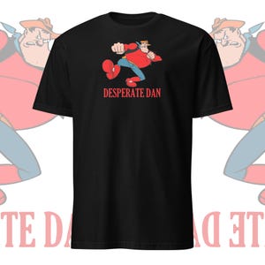 Puede incluir: Camiseta negra con una imagen de dibujos animados de Desperate Dan, un personaje de cómic, con una camisa roja y pantalones vaqueros azules. El texto "DESPERATE DAN" está impreso en rojo debajo de la imagen.