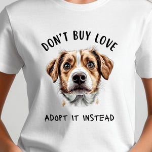 May include: White t-shirt featuring a dog illustration with the text "DON'T BUY LOVE ADOPT IT INSTEAD". The dog has brown and white fur and a sweet expression. The shirt is a classic crew neck style.