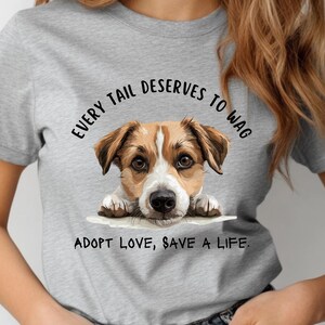 May include: A light gray t-shirt featuring a dog illustration with the text "EVERY TAIL DESERVES TO WAG" and "ADOPT LOVE, SAVE A LIFE." The dog has brown and white fur. The shirt is made of a soft material.