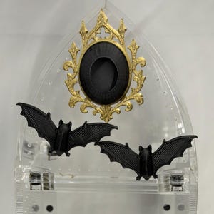 May include: A clear acrylic display stand with a pointed arch top, featuring a gold-colored ornate oval frame with a black center insert. Two black bat figures are attached to the front of the clear structure, positioned below the oval frame.