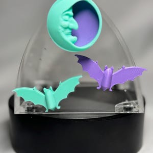 May include: A decorative Halloween-themed display featuring a teal crescent moon with a face and two bat figurines. The bats are teal and purple, set against a clear dome and black base.