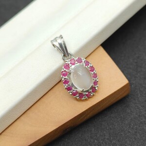 May include: A silver pendant with an oval, milky-white gemstone center, surrounded by a halo of small, vibrant red gemstones. The pendant has a simple, elegant design, suitable for everyday wear or special occasions.