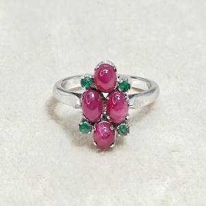 May include: A silver ring featuring a cluster of five oval, deep red gemstones, possibly rubies, surrounded by smaller green stones, likely emeralds. The ring's band is a simple, polished silver, and the gemstones are set in a floral-like arrangement.