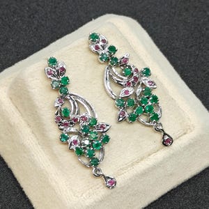 Puede incluir: Un par de pendientes colgantes plateados con un diseño floral. Cada pendiente presenta grupos de piedras preciosas verdes y rojas, con una piedra preciosa en forma de lágrima en la parte inferior. Los pendientes se exhiben sobre una superficie color crema.