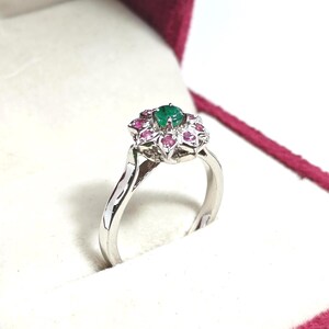 Può includere: Un anello d'argento con un design floreale. L'anello presenta una gemma verde centrale circondata da gemme rosa più piccole. L'anello è in una scatola bianca con interno bordeaux.