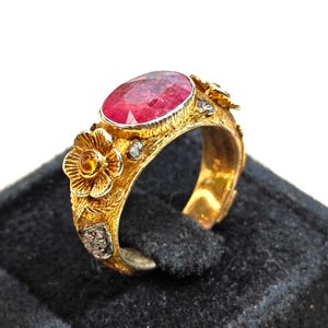 Puede incluir: Un anillo de oro con un gran rubí ovalado. El anillo está adornado con detalles florales y de hojas, y pequeñas piedras de acento. La banda tiene un acabado texturizado. El anillo se muestra sobre una superficie negra.