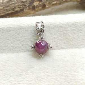 May include: A silver pendant featuring a round, deep pink gemstone. The gemstone is the focal point, set within a decorative silver setting with small, ornate details. The pendant has a loop for a chain.