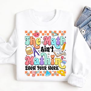 Pode incluir: Camisola branca com o texto "The Math Ain't Mathin' Show Your Work" em letras coloridas. O design inclui gráficos relacionados com a matemática, como uma calculadora, um lápis e um transferidor. A camisola tem gola redonda.