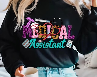 May include: Black sweatshirt with a graphic design that says "Medical Assistant" in colorful letters with a stethoscope, pills, a syringe, and a nurse's cap.