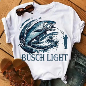 May include: White t-shirt featuring a blue and white graphic of a leaping fish, waves, and a fisherman. The text "BUSCH LIGHT" is printed in blue below the graphic. Sunglasses and sandals are also visible.