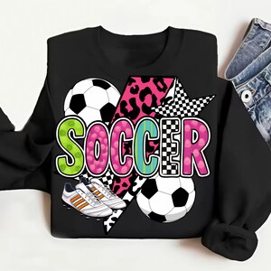Puede incluir: Sudadera negra con un dise&ntilde;o gr&aacute;fico de un bal&oacute;n de f&uacute;tbol, botas de f&uacute;tbol y la palabra "SOCCER" en letras de colores con estampado de leopardo y patr&oacute;n a cuadros.