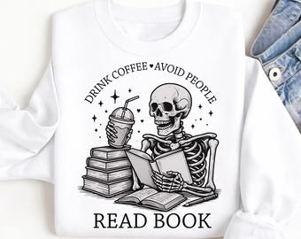 May include: White sweatshirt with a black and white graphic of a skeleton reading a book and holding a coffee cup. The text reads "DRINK COFFEE • AVOID PEOPLE" and "READ BOOK". The design includes stars and a pen.