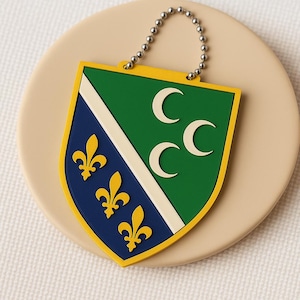 Peut inclure: Un porte-clés en forme de bouclier avec un dessin d'armoiries. Le bouclier a une section supérieure verte avec trois croissants de lune blancs, une section inférieure bleue avec trois fleurs de lys dorées et une bande diagonale blanche. Une chaîne à billes argentée est attachée.