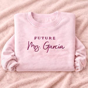 May include: A light pink sweatshirt with the words "FUTURE Mrs. Garcia" embroidered in a darker pink script. The sweatshirt is folded and resting on a pink fabric surface. The design is a bridal theme.