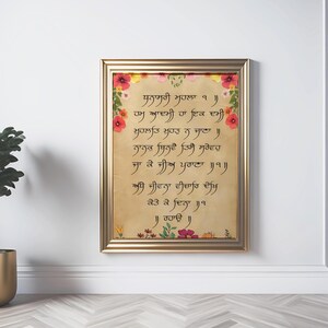 May include: A framed wall art piece featuring Gurmukhi script printed on a parchment-colored background. The text is surrounded by a decorative border of colorful red, yellow, and pink flowers and green leaves, all encased in a metallic gold frame.