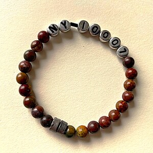 Può includere: Un braccialetto di perline marroni e rosse con perline d'argento che compongono la scritta "NY 10007".