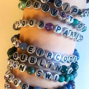 Puede incluir: Un conjunto de seis pulseras con cuentas plateadas y letras negras que deletrean diferentes palabras y números. Las pulseras están hechas con piedras de diferentes colores, incluyendo verde, azul, morado y marrón.