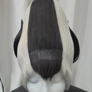 May include: A wig with a black and white color scheme, styled on a mannequin head. The wig features a central black stripe flanked by white sections, with black ear-like extensions on each side. The wig is likely for cosplay or costume use.