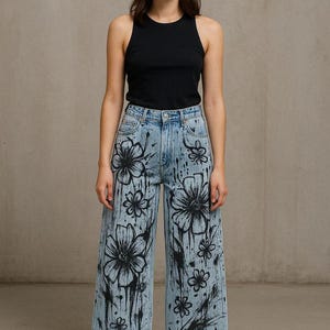 K&ouml;nnte beinhalten: Ein schwarzes Tanktop kombiniert mit hellblauen Jeans mit gro&szlig;en, schwarzen Blumendesigns. Die Jeans haben einen weiten Beinverlauf und sind mit farb&auml;hnlichen Spritzern verziert, was einen einzigartigen, k&uuml;nstlerischen Look erzeugt. Schwarze Schuhe vervollst&auml;ndigen das Outfit.
