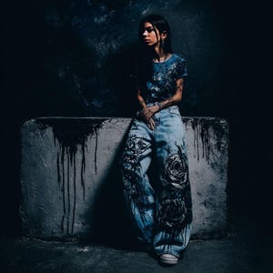 K&ouml;nnte beinhalten: Eine Person tr&auml;gt ein blaues T-Shirt und eine hell gewaschene Jeans mit gro&szlig;en schwarzen Rosendesigns. Die Jeans hat eine weite Beinpassform. Der Hintergrund ist dunkel mit einem Betonblock.