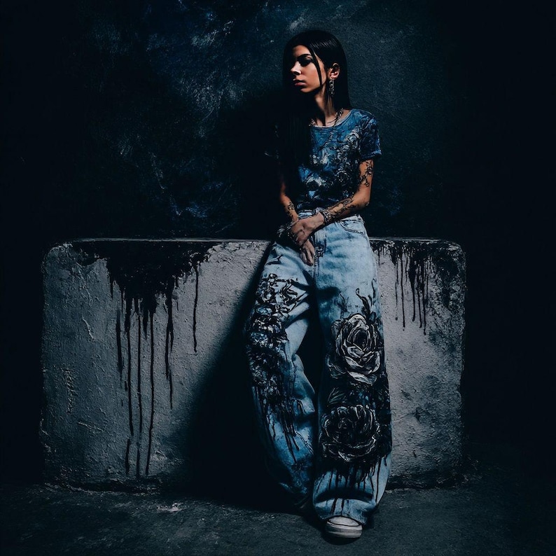 K&ouml;nnte beinhalten: Eine Person tr&auml;gt ein blaues T-Shirt und eine hell gewaschene Jeans mit gro&szlig;en schwarzen Rosendesigns. Die Jeans hat eine weite Beinpassform. Der Hintergrund ist dunkel mit einem Betonblock.
