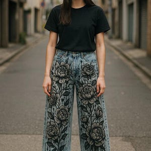 K&ouml;nnte beinhalten: Hellblaue Jeans mit gro&szlig;em Blumenmuster in Schwarz und Grau. Die Jeans hat einen weiten Bein-Schnitt und wird mit einem schwarzen T-Shirt und einer M&uuml;tze getragen.