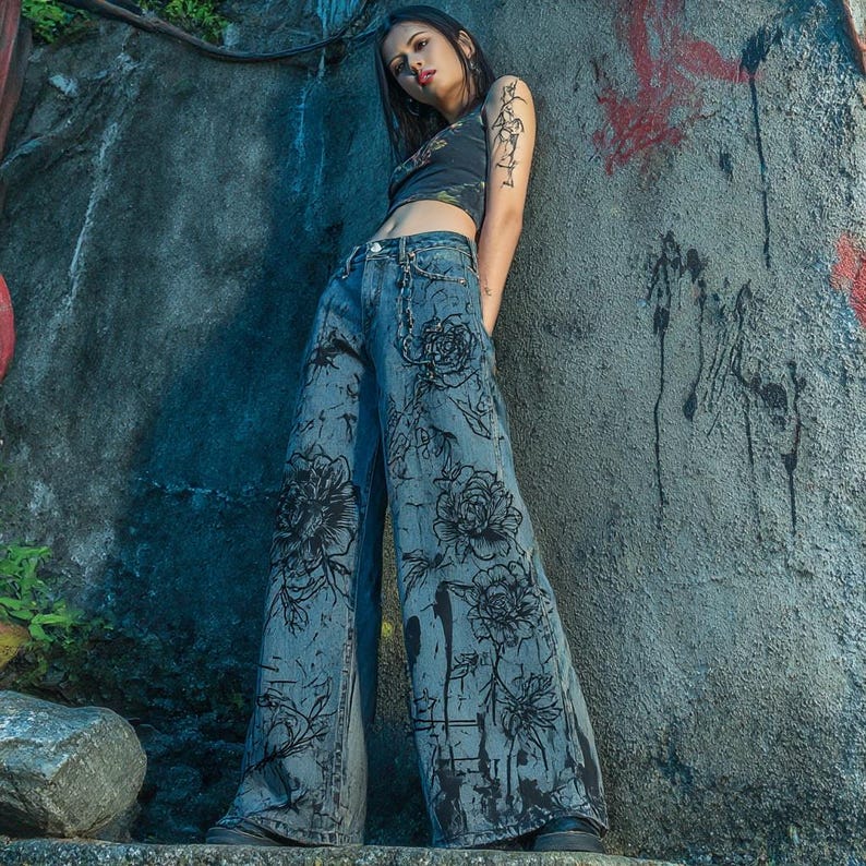 K&ouml;nnte beinhalten: Weit geschnittene blaue Jeans mit einem Blumendesign in schwarzer Tinte. Die Jeans haben eine hohe Taille und werden mit einem dunklen Crop-Top kombiniert. Das Model lehnt an einer strukturierten grauen Wand.