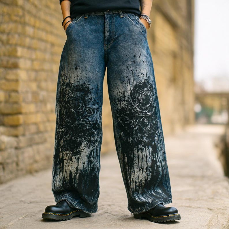 K&ouml;nnte beinhalten: Blaue Jeans mit weitem Bein und einem schwarzen Rosendesign sowie Farbspritzern. Die Jeans werden mit schwarzen Stiefeln getragen. Die Jeans haben einen Vintage-Look.