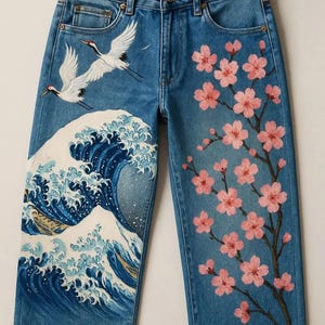 May include: Blue denim jeans with hand-painted artwork. The design features a large ocean wave, two flying cranes, and a flowering cherry blossom branch. The jeans have a classic five-pocket design.