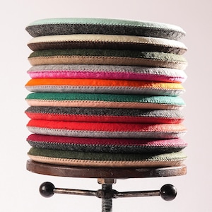 Könnte beinhalten: Ein Stapel aus zwölf runden Sitzkissen aus Filz in verschiedenen Farben wie Minzgrün, Creme, Dunkelgrün, Beige, Pink, Orange, Türkis, Rot, Grau und Bordeaux, die auf einem verstellbaren Hocker aus dunklem Metall mit einer Gewindestange liegen.