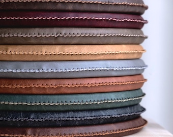Puede incluir: Una pila de cojines redondos y planos con una superficie lisa de textura mate en varios colores, incluyendo marrón oscuro, burdeos, beige, gris claro, terracota, verde bosque y negro, cada uno con un borde acabado en punto de festón.