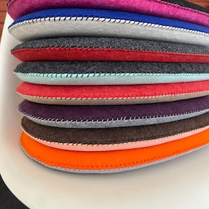 May include: A stack of nine round felt chair cushions in various colors, including pink, blue, gray, red, teal, beige, purple, brown, and orange. Each cushion has a contrasting color trim around the edge.