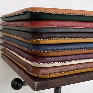 May include: A stack of rectangular leather sleeves in various colours including tan, dark green, red, black, yellow, navy blue, brown, burgundy, and cream. Each sleeve features visible hand-stitched edges with contrasting thread along the perimeter.