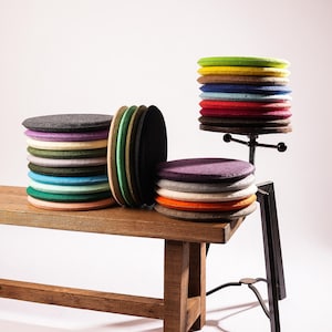 Op de afbeelding: Een collectie ronde vilten zitkussens in diverse kleuren, waaronder groen, geel, bruin, blauw, rood, paars, oranje en grijs. De kussens zijn gerangschikt in drie stapels, waarvan één op een houten bank en een andere op een metalen standaard.