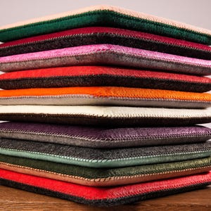 Puede incluir: Una pila vertical de once fundas rectangulares de fieltro en varios colores, incluyendo melocotón, verde oscuro, burdeos, rosa, rojo, naranja, crema, morado, gris, verde claro y oliva, cada una con bordes cosidos visibles en el perímetro.