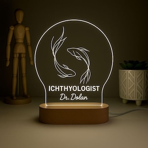 May include: An illuminated LED lamp with a wooden base. The lamp has a clear acrylic panel with a white outline of two koi fish and the text "ICHTHYOLOGIST Dr. Dolan". The lamp is designed to provide a warm glow.