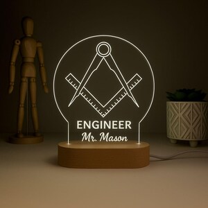 Puede incluir: Lámpara LED iluminada con un símbolo masónico, con un diseño de escuadra y compás, y el texto "ENGINEER Mr. Mason". La lámpara tiene una base de madera y un panel de luz circular transparente. El diseño general es una luz cálida y acogedora.