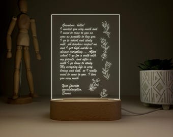 Könnte beinhalten: Eine beleuchtete Acrylplatte mit Holzsockel zeigt eine herzliche Botschaft an eine Großmutter, mit floralen Akzenten. Die Botschaft lautet: "Oma, hallo! Ich habe dich sehr vermisst... Ich liebe dich sehr. Deine Lieblingsenkelin, Emma."