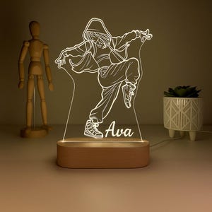 May include: A personalized LED night light featuring a dancer in a dynamic pose. The design is etched into a clear acrylic panel, illuminated by a warm light from a wooden base. The name "Ava" is displayed below the dancer.