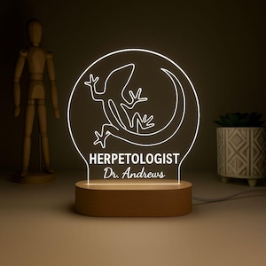May include: A glowing LED night light with a reptile design. The light features a circular design with a lizard illustration and the text "HERPETOLOGIST Dr. Andrews" on a wooden base.