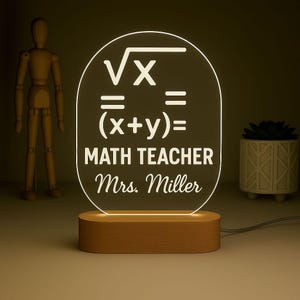 Puede incluir: Lámpara de escritorio LED con base de madera. El panel acrílico transparente muestra símbolos matemáticos y el texto "MATH TEACHER Mrs. Miller". La lámpara emite una luz cálida y acogedora.