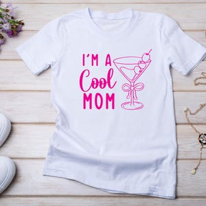 Pu&ograve; includere: T-shirt bianca con la scritta rosa "I'M A Cool MOM" e un'illustrazione di bicchiere da Martini rosa con olive e un fiocco. La maglietta &egrave; su una superficie chiara effetto legno.