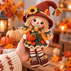 May include: A crocheted scarecrow doll wearing a dark orange pointed hat with a sunflower and leaf, a yellow long-sleeve shirt, and a dark green overall dress. The doll has blonde yarn hair in braids, holds a small orange pumpkin, and wears brown boots.