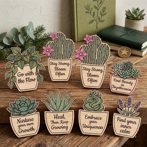 Op de afbeelding: Acht houten plantenbordjes met gedrukte tekst. Elk bordje toont een andere plant, waaronder cactussen, vetplanten en lavendel, in lichtgekleurde houten potten. De tekst bevat zinnen zoals Go with the Flow, Stay Strong, Bloom Often en Find your inner calm.