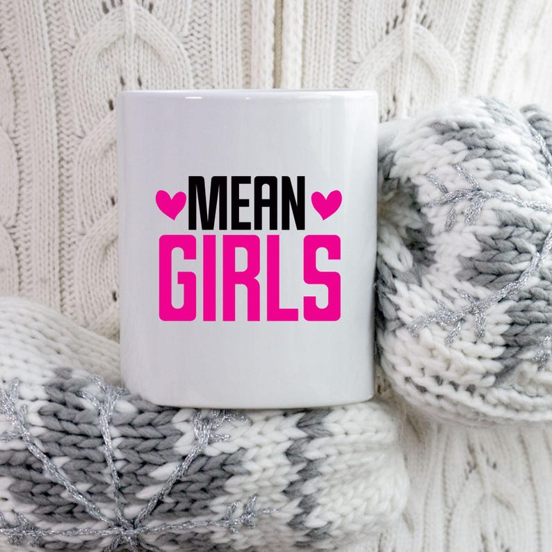 Pu&ograve; includere: Una tazza di ceramica bianca con le parole "MEAN GIRLS" in lettere nere e rosa. La tazza &egrave; circondata da un maglione e guanti di lana bianchi e grigi.