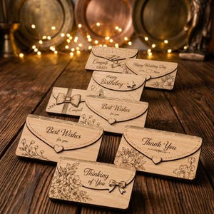 Puede incluir: Una colección de sobres rectangulares de madera con cierre de solapa, cada uno con textos e ilustraciones grabados con láser. Las frases incluyen Congrats Grad, Happy Wedding Day, Happy Birthday, Best Wishes y Thank You, decoradas con motivos florales.