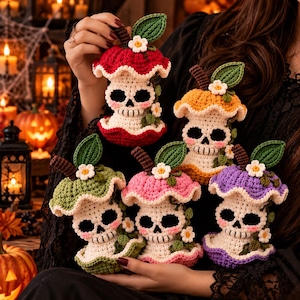 May include: Five crocheted skull figures with mushroom-shaped caps in red, orange, green, pink, and purple. Each skull features black eye sockets, stitched teeth, pink cheeks, a brown stem, a green leaf, and a small white flower on top.