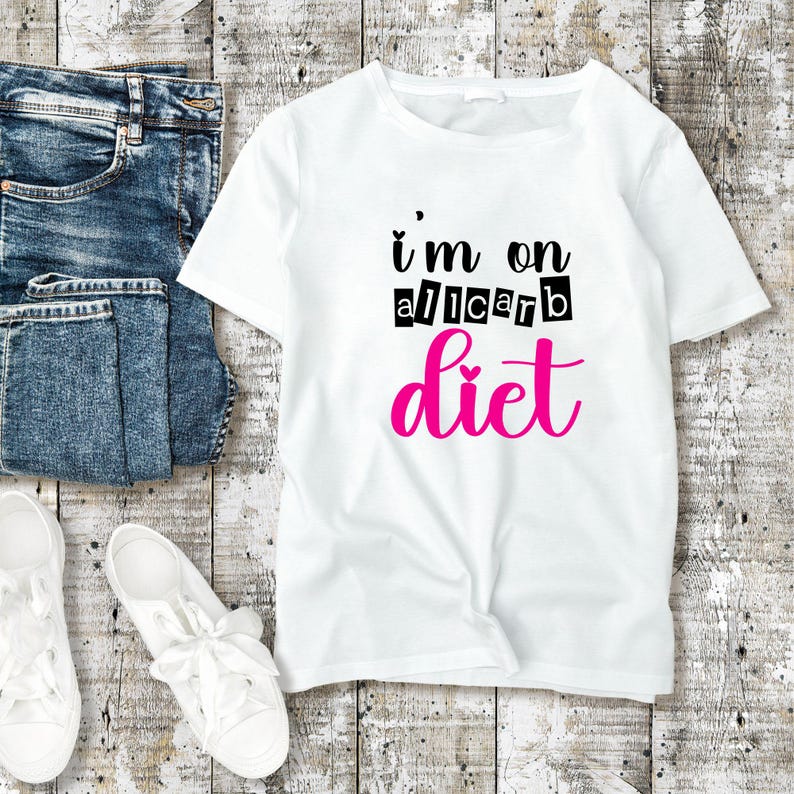 Pu&ograve; includere: T-shirt bianco con la scritta "I'm on a carb diet" in nero e rosa. La maglietta &egrave; abbinata a jeans blu e scarpe da ginnastica bianche su uno sfondo di legno.