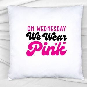 Pu&ograve; includere: Cuscino quadrato bianco con la scritta "ON WEDNESDAY We Wear Pink" in lettere nere e rosa. Il testo rosa include piccoli cuori. Il cuscino &egrave; su una superficie bianca.